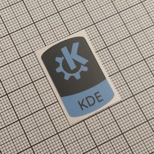 Peut inclure: Un autocollant avec un design noir et bleu. La partie supérieure est noire avec un engrenage bleu et la lettre "K". La partie inférieure est bleue avec les lettres "KDE". L'autocollant est sur un fond blanc avec un motif quadrillé.