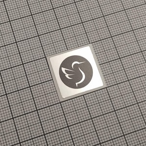 May include: A square sticker with a silver border and a gray background. The sticker features a circular design with a white bird silhouette. The bird has a long beak and a curved neck, with a wing extended.