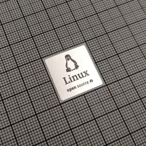 Peut inclure: Un autocollant carré argenté avec le mot "Linux" et un logo de pingouin. Le texte "open source" est en dessous. L'autocollant est posé sur un fond de grille grise.
