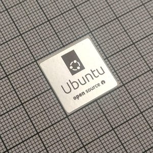 Könnte beinhalten: Ein quadratischer silberner Aufkleber mit dem Ubuntu-Logo und Text. Das Logo ist ein Kreis aus miteinander verbundenen Punkten über dem Wort "Ubuntu" und dem Text "open source". Der Aufkleber befindet sich auf einem grauen Gitterhintergrund.