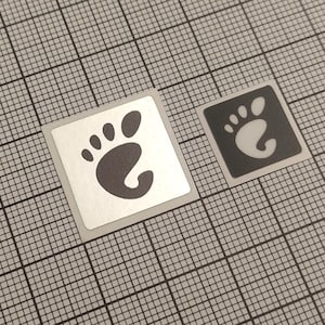 Op de afbeelding: Twee vierkante stickers met een zwart voetprintontwerp. Eén sticker heeft een zilveren achtergrond, de andere een zwarte achtergrond. De stickers zijn geplaatst op een grijze rasterachtergrond.