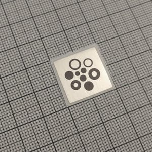Könnte beinhalten: Ein quadratischer, silberner Aufkleber mit einem schwarzen kreisförmigen Design. Das Design besteht aus unterschiedlich großen Kreisen und Ringen, die in einer Anordnung angeordnet sind. Der Aufkleber befindet sich auf einem grauen Gitterhintergrund.