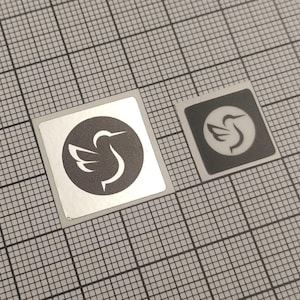 May include: Two square stickers with a bird logo. One sticker is silver with a black circle and a white bird design. The other sticker is smaller, black with a white bird design. The stickers are on a gray grid.