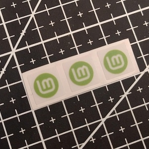 May include: Three small square stickers arranged in a horizontal row. Each sticker features a circular light green background with a white stylized letter M logo centered inside. The stickers have a thin white border around the circular design.