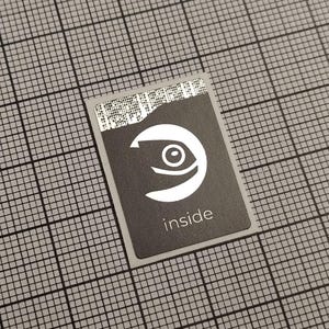 Pode incluir: Um autocolante retangular cinzento com um logótipo branco de um olho dentro de uma forma de crescente e a palavra "inside" por baixo. A borda superior tem um código de barras parcialmente rasgado. O autocolante está num fundo de grelha cinzenta.