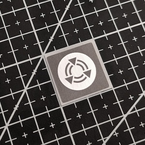 Könnte beinhalten: Ein kleiner, quadratischer, grauer Aufkleber mit einem weißen kreisförmigen Grafikdesign mit drei nach innen zeigenden Pfeilen. Der Aufkleber befindet sich auf einer schwarzen Schneidematte mit einem Rastermuster aus weißen Linien und Pluszeichen.