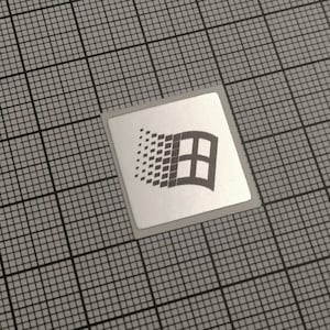 Könnte beinhalten: Ein quadratischer Aufkleber mit silbernem Rand, der das Windows-Logo zeigt. Das Logo ist eine stilisierte Flagge mit einem gitterartigen Muster auf der linken Seite und einer Fensterform auf der rechten Seite. Der Aufkleber befindet sich auf einem grauen Rasterhintergrund.