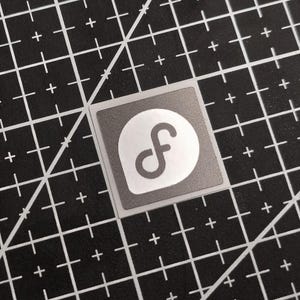 May include: A square sticker with a gray border and a white circle containing a stylized, lowercase "f" in gray. The sticker is on a black cutting mat with white grid lines.