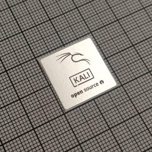 May include: A square, silver sticker with the word "KALI" in a black box, and the words "open source" below it. A stylized black logo is above the word "KALI". The sticker is on a gray grid background.