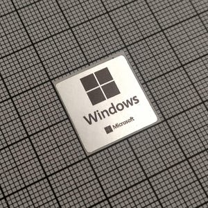 Könnte beinhalten: Ein silberner quadratischer Aufkleber mit dem Windows-Logo, einem Design mit vier Fenstern und dem Wort "Windows" in einer serifenlosen Schriftart. Das Microsoft-Logo befindet sich unter dem Wort "Windows". Der Aufkleber befindet sich auf einem grauen Raster.