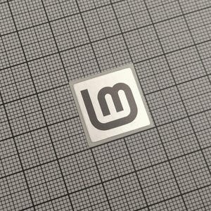 May include: A square sticker with a silver border and a grey logo featuring the letters "lm" in a stylized design. The sticker is set against a grey grid background.