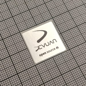 May include: A square, silver sticker with the text "XVUAN" in a stylized font and a curved arrow design. Below the logo, the words "open source" are printed in a smaller font. The sticker is on a gray grid background.
