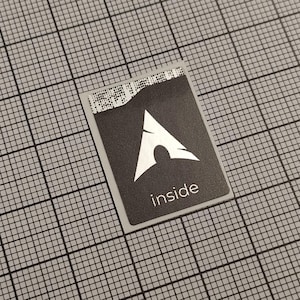 Peut inclure: Un autocollant rectangulaire avec un fond noir et un logo blanc représentant une flèche pointant vers le haut avec une base incurvée. Le mot "inside" est imprimé en blanc en dessous. L'autocollant est sur une grille grise.