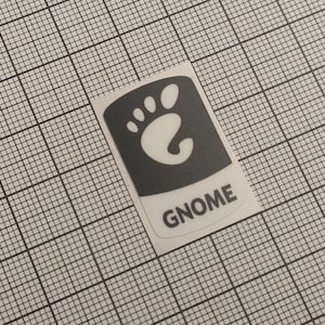 Op de afbeelding: Een sticker met het Gnome-logo, een gestileerde voetafdruk in een donkergrijze, afgeronde rechthoek. Het woord "GNOME" staat eronder in een schreefloze lettertype. De sticker staat op een rasterachtergrond.