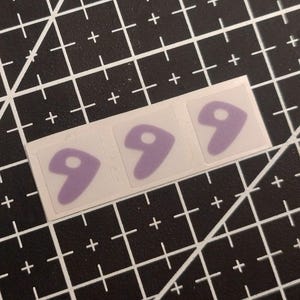 Puede incluir: Tres pegatinas individuales dispuestas en una fila horizontal. Cada pegatina presenta una forma abstracta de color morado claro similar a una coma con un pequeño recorte circular cerca de la parte superior, sobre una lámina transparente.