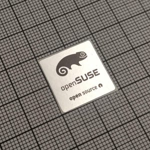 May include: A square, silver sticker with the text "openSUSE open source" and a stylized chameleon logo. The sticker is on a gray grid background.