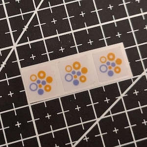 May include: A strip of three square stickers, each featuring a cluster of small circles in shades of orange and light blue. The stickers are arranged horizontally, displaying a repeating pattern of solid and outlined circular shapes on a white background.