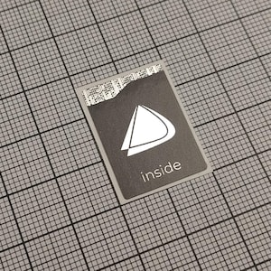 Könnte beinhalten: Ein rechteckiger, dunkelgrauer Aufkleber mit einem weißen geometrischen Design und dem Wort "inside" in Weiß gedruckt. Der Aufkleber hat eine zerrissene Kante oben und befindet sich auf einem grauen Rasterhintergrund.