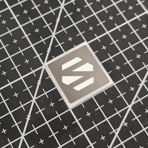 Könnte beinhalten: Ein quadratischer Aufkleber mit grauem Hintergrund und weißem geometrischem Design. Der Aufkleber liegt auf einer schwarzen Schneidematte mit weißem Rastermuster.