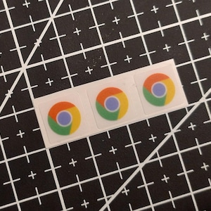Könnte beinhalten: Drei kleine, quadratische Aufkleber in einer horizontalen Reihe, die jeweils das kreisförmige Google Chrome-Logo mit seinen charakteristischen roten, gelben, grünen und blauen Farbsegmenten auf weißem Grund zeigen.