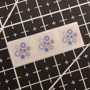 May include: Three square stickers arranged in a horizontal row, each featuring a cluster of small blue and grey circles of varying sizes. The stickers are translucent with a white border and are displayed on a black grid cutting mat.
