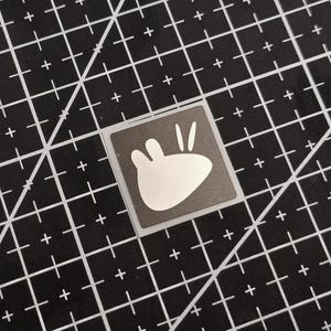 May include: A square sticker featuring a white cartoon rabbit design on a dark grey background. The sticker is on a black cutting mat with white grid lines and diagonal lines.