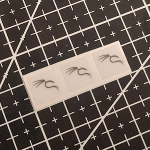 Könnte beinhalten: Drei kleine, quadratische, durchscheinende Aufkleber in einer horizontalen Reihe, jeder mit einer schwarzen, minimalistischen Strichzeichnung eines Drachenkopfprofils mit geschwungenen Hörnern oder Schnurrhaaren auf einer schwarzen Schneidematte.