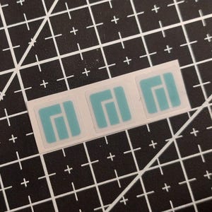 May include: Three square stickers arranged in a horizontal row on a white backing strip. Each sticker features a light blue graphic design consisting of a solid square with a vertical bar and a shorter adjacent bar, creating a geometric pattern.