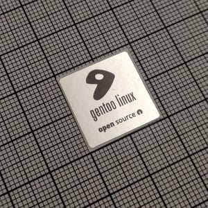 May include: A square, silver sticker with the text "gentoo linux open source" and a stylized black logo. The sticker is centered on a grey grid background.