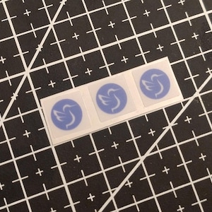 Puede incluir: Tres pegatinas cuadradas dispuestas en una fila horizontal, cada una con un fondo circular azul claro y una silueta de pájaro estilizada de color blanco en el centro. Las pegatinas se presentan sobre una lámina de soporte transparente.