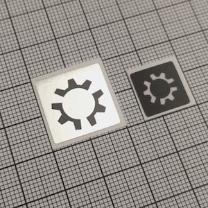 May include: Two square stickers with a gear design. One sticker has a silver background with a black gear, and the other has a black background with a white gear. The stickers are on a gray grid background.