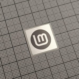Può includere: Un adesivo quadrato con bordo argentato con un cerchio grigio con un logo "lm" bianco. L'adesivo è centrato su uno sfondo a griglia grigia.