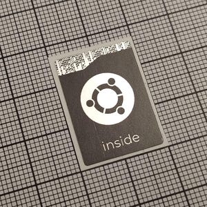 Pode incluir: Um pequeno cartão retangular com fundo preto e logotipo Ubuntu branco. A palavra "inside" está impressa em branco abaixo do logotipo. O cartão está sobre um fundo de grade cinza.