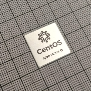 May include: A square, silver sticker with the word "CentOS" in a bold, sans-serif font. Below it, the text "open source" is displayed. The sticker features a geometric design above the word "CentOS" and is set against a grid background.