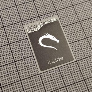Pode incluir: Um autocolante retangular com fundo cinza escuro e um logótipo de dragão branco. A palavra "inside" está impressa em branco na parte inferior. O autocolante está numa superfície de grelha cinzenta.