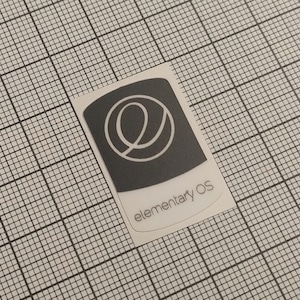 Puede incluir: Una pegatina rectangular con fondo gris oscuro y un logotipo 'e' blanco estilizado. El texto 'elementary OS' está impreso en gris claro debajo. La pegatina está sobre un fondo de cuadrícula.