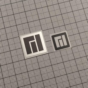 May include: Two square stickers with a geometric design. The larger sticker has a silver border and a black and white graphic. The smaller sticker has a black and white graphic. Both stickers are on a gray grid background.