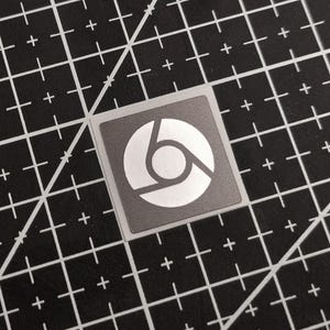 May include: A square sticker with the Chrome logo, featuring a white, stylized gear-like design on a gray background. The sticker is placed on a black cutting mat with white grid lines.