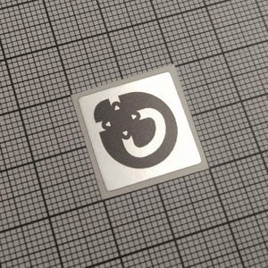 May include: A square sticker with a silver border and a gray design. The design is a stylized letter or symbol, featuring a curved shape with cutouts. The sticker is on a gray grid background.