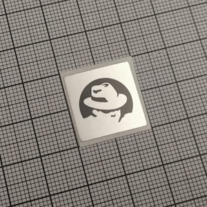 May include: A square sticker with a silver border features a black and white illustration of a person wearing a fedora hat. The sticker is placed on a gray grid background.