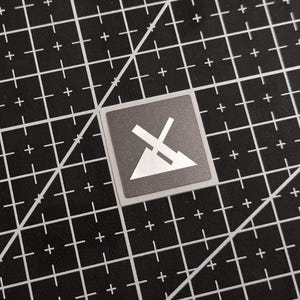 Könnte beinhalten: Ein quadratischer, grauer Aufkleber mit einer weißen Grafik eines stilisierten Berges und einem X. Der Aufkleber befindet sich auf einer schwarzen Schneidematte mit weißen Rasterlinien und diagonalen Linien.
