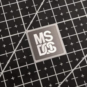 Op de afbeelding: Een vierkante sticker met de letters "MS" boven "DS" in een witte, gestileerde letter. De sticker ligt op een zwarte snijmat met witte rasterlijnen.
