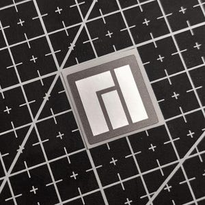 May include: A square, silver sticker with a gray border and a white geometric design. The sticker is on a black cutting mat with white grid lines and plus signs.