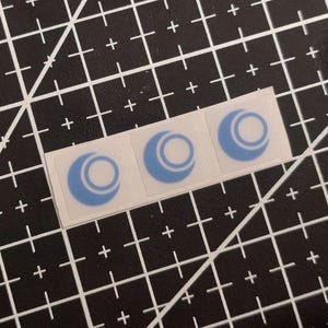 Peut inclure: Trois autocollants carrés disposés en rangée horizontale, chacun présentant une forme de croissant de lune bleu clair entourant partiellement un cercle blanc uni au centre. Les autocollants ont un support adhésif transparent sur une grille noire.
