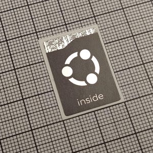 Könnte beinhalten: Eine rechteckige, dunkelgraue Karte mit einer weißen, kreisförmigen Grafik und dem Wort "inside" in Weiß gedruckt. Die Karte hat einen silbernen Rand und liegt vor einem Rasterhintergrund.