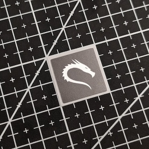 May include: A square sticker with a white dragon logo on a gray background. The sticker is on a black cutting mat with white grid lines.