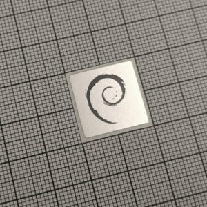 Puede incluir: Una pequeña pegatina cuadrada con un borde plateado y un diseño de espiral negro estilizado. La pegatina está sobre un fondo de cuadrícula gris, creando un efecto visual contrastante. La espiral recuerda a una pincelada.