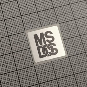 Op de afbeelding: Een vierkante sticker met de letters "MS DOS" in een vet, schreefloos lettertype. De letters zijn donkergrijs op een lichtgrijze achtergrond. De sticker staat op een grijze rasterachtergrond.