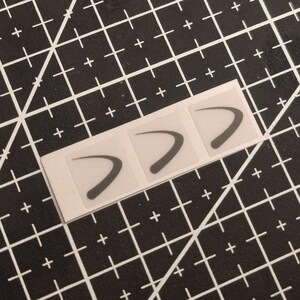 May include: A rectangular strip of white backing paper containing three individual square stickers. Each sticker features a dark gray, curved, boomerang-shaped graphic design centered within the square border on a translucent background.
