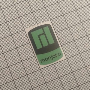 Peut inclure: Un autocollant rectangulaire avec le logo Manjaro. Le logo est composé de formes vertes et noires avec le mot "manjaro" en vert en dessous. L'autocollant est posé sur un fond quadrillé.